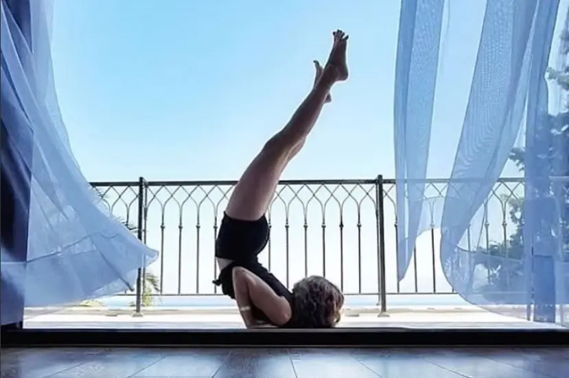 A shoulder stand backbend on a balcony between sheer blue curtains, with the sea beyond