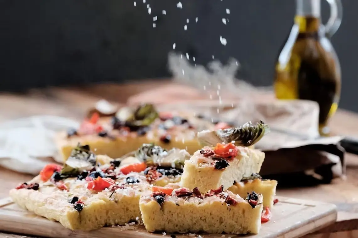 Focaccia topped with tomato, olives and artichoke, with a bottle of olive oil behind
